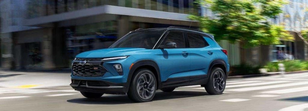 A light blue SUV with a black roof drives on a city street with buildings and trees in the blurred background.