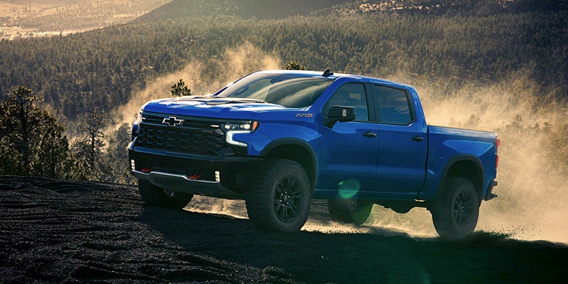 Blue pickup truck driving across a dusty hilltop trail, kicking up clouds of dirt