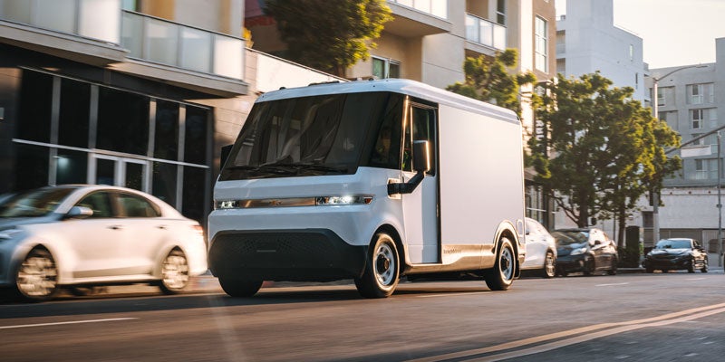 A white electric delivery van drives down a city street with blurred background cars and buildings.