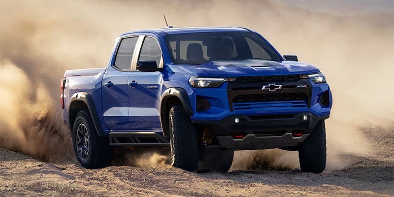 Blue pickup truck driving over sandy off-road terrain, kicking up thick clouds of dust behind it