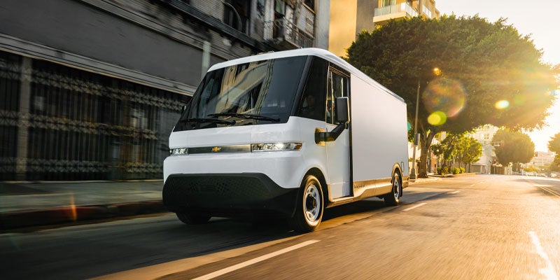 A white Chevrolet BrightDrop delivery van with a large windshield drives on a city street toward a bright sunset.