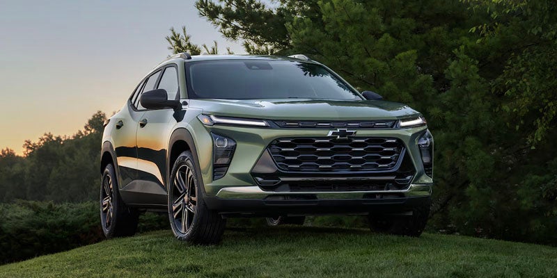 Front view of a dark green Chevrolet Trax Activ SUV parked on a grassy hill with green trees in the background.