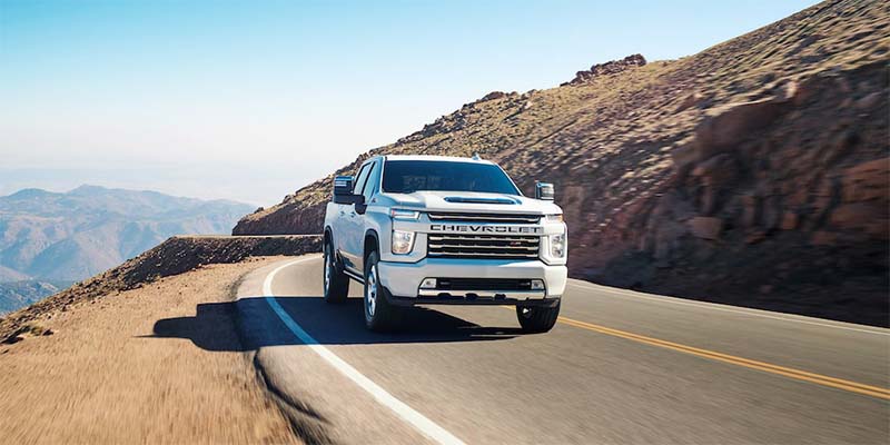 White Chevrolet pickup truck drives on a winding mountain road with distant peaks under a clear sky.