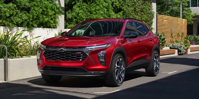 A shiny red 2024 Chevrolet Trax RS parked diagonally in a lot, surrounded by lush green foliage and sun