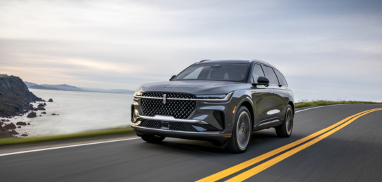 A sleek, gray SUV drives on a coastal highway with a scenic ocean view on its left. The sky is overcast, conveying a calm, adventurous tone.