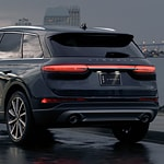 A sleek, dark gray SUV is parked on a wet street at dusk. The vehicle's rear lights glow softly, and a city skyline is visible in the background.