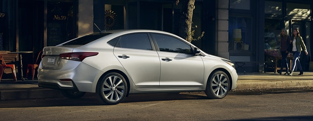 A sleek silver sedan is parked on a sunlit street, casting shadows. Two people walk on the sidewalk beside a cafe. Urban and modern tone.