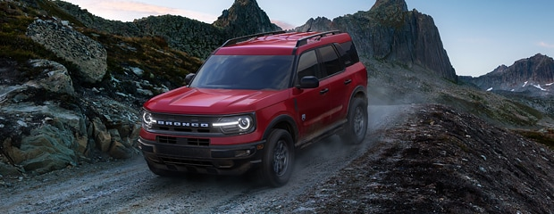 A red SUV drives on a rugged mountain path under a twilight sky. Dust clouds trail behind, conveying adventure and ruggedness. Jagged peaks loom in the background.