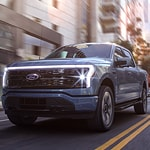 A sleek, modern truck drives down a city street lined with tall buildings. The vehicle's LED headlights are on, capturing a dynamic, urban vibe.