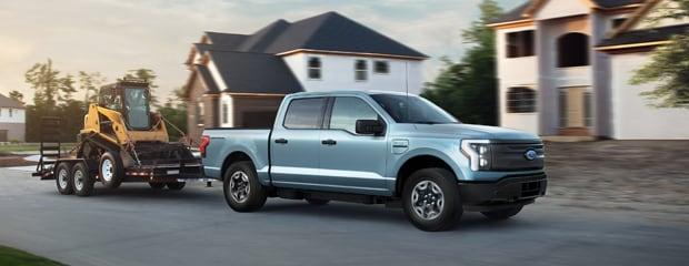 A blue pickup truck tows a yellow construction vehicle on a trailer past large suburban homes. The scene conveys strength and utility.