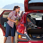 A woman in outdoor attire stands by an open red SUV, organizing climbing gear. The scene is set in a sunny, mountainous area, conveying adventure.
