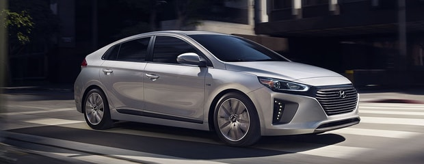 Silver hatchback car driving on a sunlit city street. The vehicle exhibits sleek, modern lines with a prominent grille, evoking a sense of sophistication.