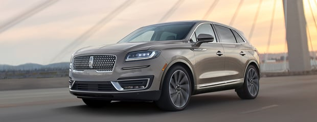 An elegant SUV, gray in color, drives on a bridge at sunset. The vehicle's sleek design features modern headlights and a polished grille, conveying luxury.