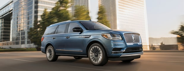 A sleek blue SUV is driving on a city street with modern glass skyscrapers in the background, conveying luxury and urban sophistication.