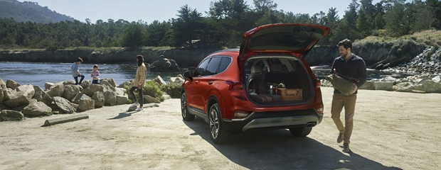 Red SUV parked near a scenic river, trunk open, revealing picnic supplies. People relax by the water on boulders. The scene is peaceful and inviting.