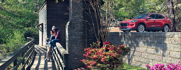 A red SUV is parked on a stone wall near a rustic building. Two people sit on a wooden bridge, surrounded by lush greenery and blooming flowers.