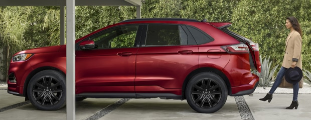 A woman in a beige coat and black boots walks toward a red SUV parked under a carport. The setting is outdoors, with lush greenery in the background.