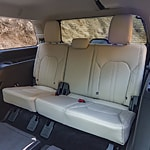 Rear view of a car interior featuring beige leather seats with black seatbelts. The window reveals a rocky, sunlit landscape outside.