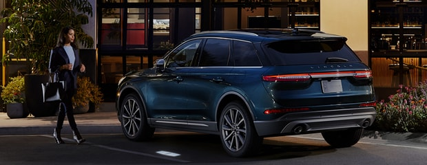 A woman walks confidently towards a parked, blue SUV outside a modern building at dusk. The scene conveys sophistication and urban elegance.
