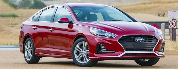 A sleek, red sedan with a modern design is parked on a road. The car is angled slightly to the left, against a background of dry, grassy hills.