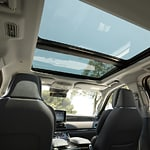 Car interior featuring two black leather seats, large panoramic sunroof showing a clear blue sky and trees, conveying openness and luxury.