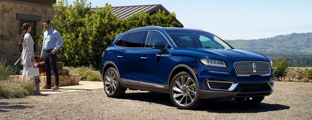 A sleek blue SUV is parked on a gravel driveway near a stone house. A man and woman stand nearby, chatting. The scene is serene with lush hills in the background.