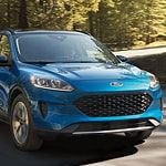 A blue SUV drives on a sunlit forest road, surrounded by tall trees. The scene conveys a sense of adventure and freedom in nature.