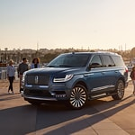 A sleek, blue SUV is parked on a sunlit street, with people walking in the background. The scene conveys a sense of modern luxury and urban lifestyle.