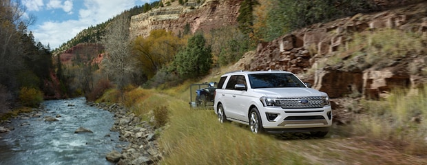 A white SUV tows a boat along a riverside trail, surrounded by rugged cliffs and lush trees. The scene conveys adventure and natural exploration.