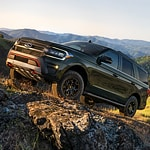 A rugged SUV navigates rocky terrain with mountains in the distance under a clear sky, conveying adventure and resilience.
