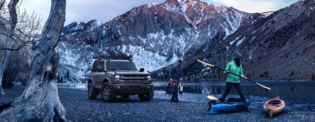 SUV parked by a serene lake with snow-capped mountains. Two people prepare kayaks, exuding a sense of adventure and tranquility amidst nature.