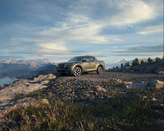 A rugged pickup truck is parked on a rocky mountain terrain under a vast, cloudy sky, set against a backdrop of snow-capped peaks and distant trees.
