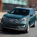 A green SUV drives on a city street. The vehicle's front grille and sleek design reflect urban sophistication, suggesting movement and modernity.