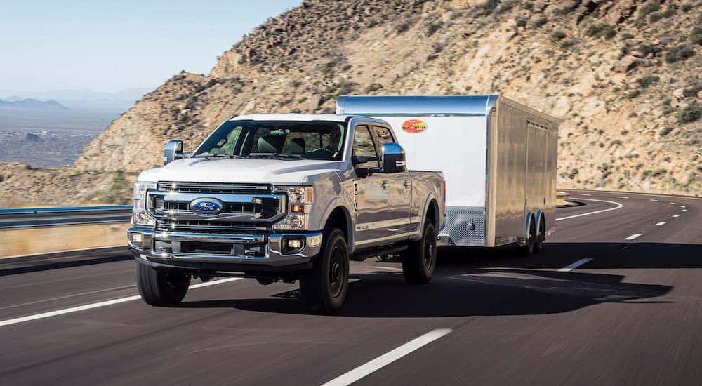 A white 2020 Ford F-350 is towing a white enclosed trailer on a highway past hills.