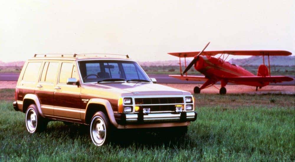 A brown 1984 Jeep Wagoneer is parked in front of a red biplane.