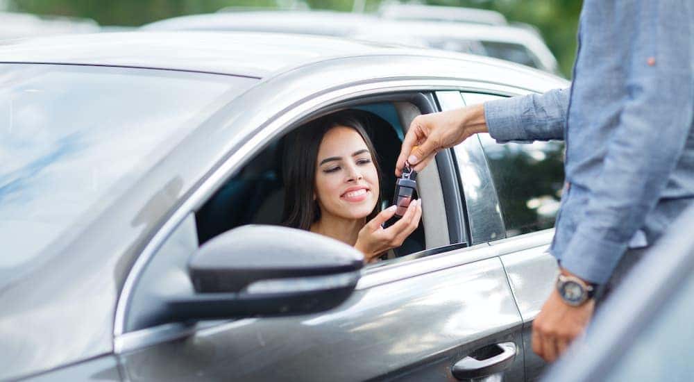 A woman is sitting in the drivers seat of a car while a salesman passes her a set of keys.