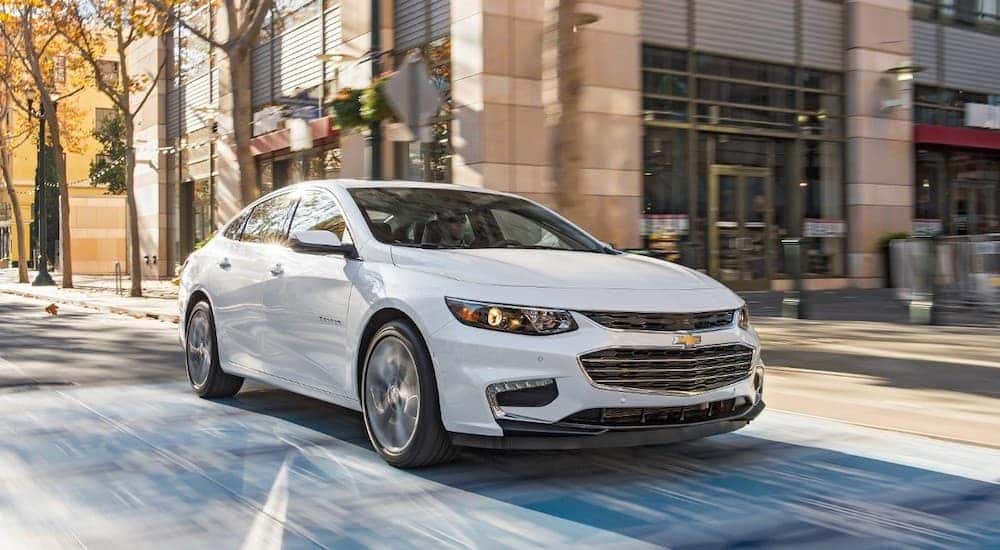 A white 2018 Chevy Malibu is driving through a city intersection.