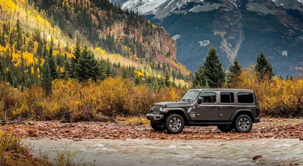 A grey 2018 Jeep Wrangler Unlimited is parked off-road in front of a river and mountains.