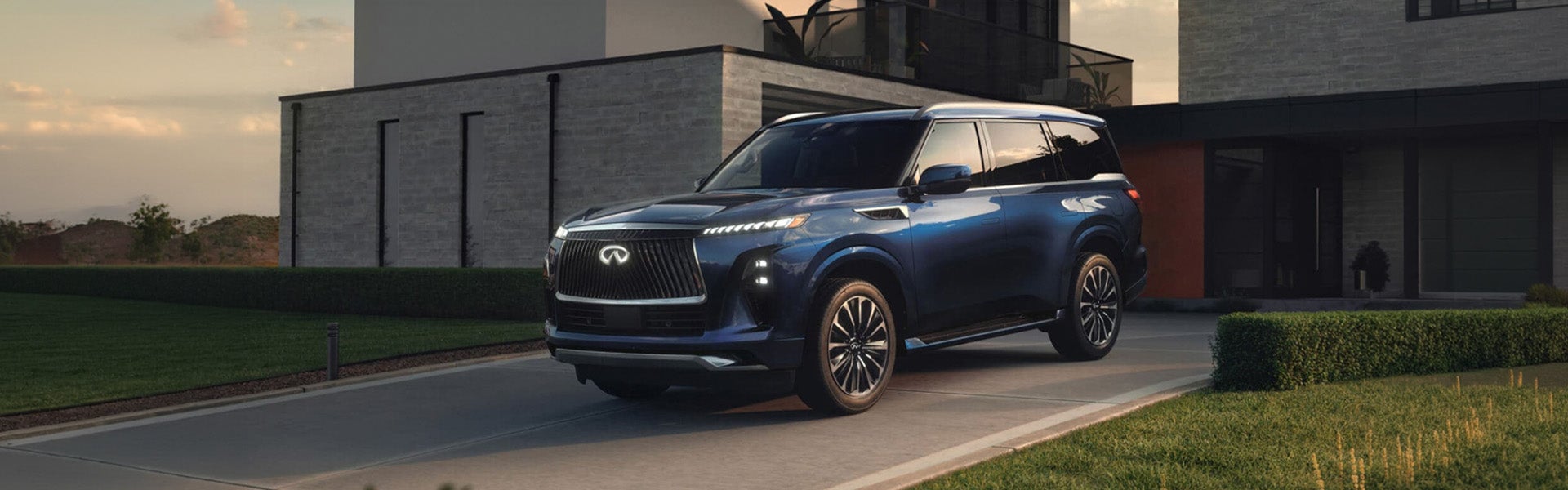 A blue 2026 INFINITI QX80 SUV parked in a modern driveway next to a stylish house at sunset.