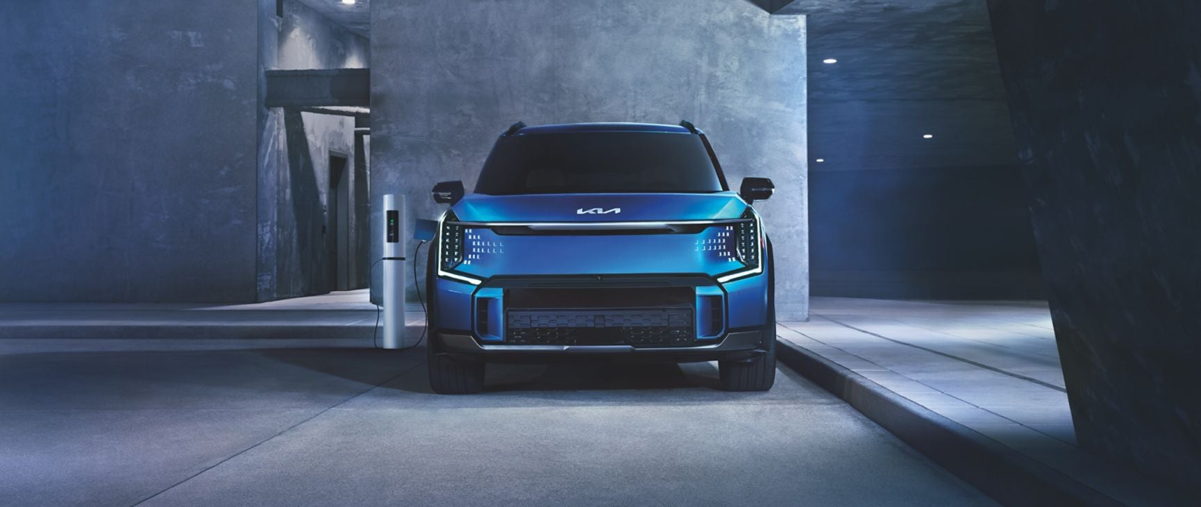 A futuristic blue Kia EV9 electric SUV is parked next to a charging station in an urban setting