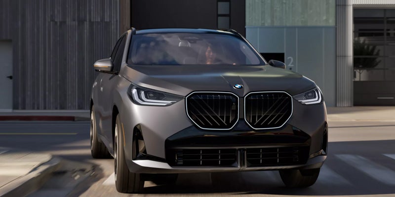 Sleek gray BMW X3 driving on a city street, showcasing its modern design and bold front grille. The scene conveys a sense of power and elegance.