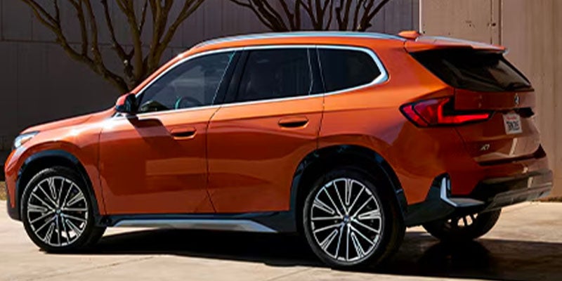 A modern orange 2025 BMW X1 SUV parked with trees in the background and a clear sky.