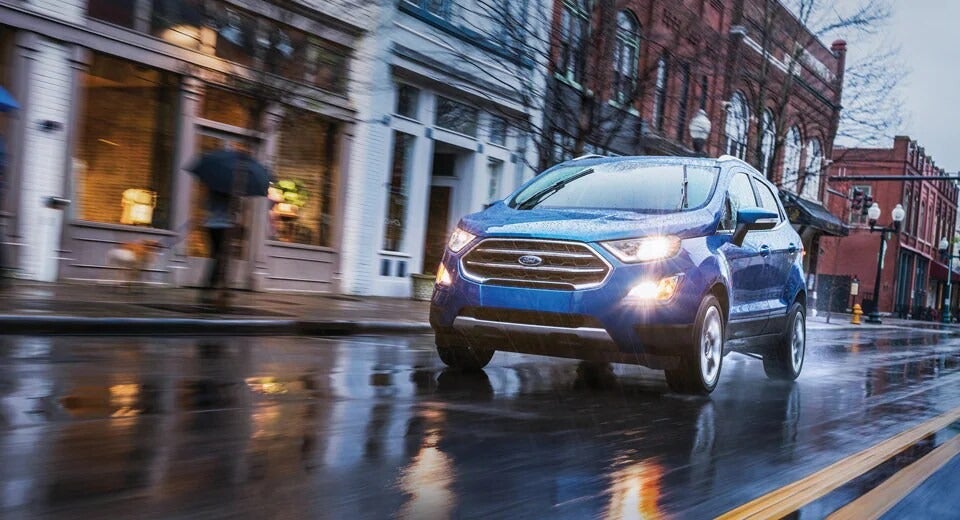 blue compact Ford driving in the rain with headlights on in a small downtown