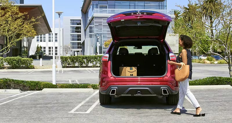 Ford SUV with open trunk in parking lot, person loading Amazon package near modern building with landscaping