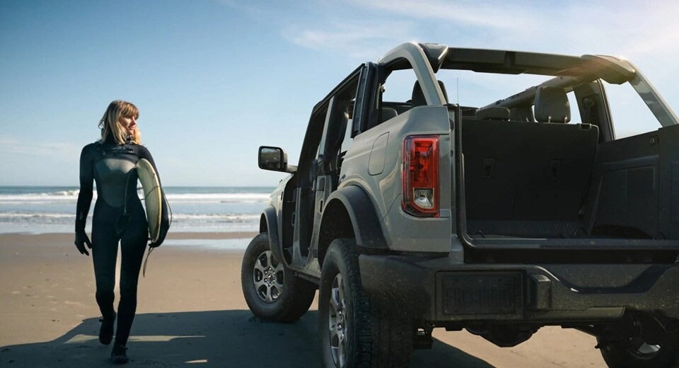 surfer walking from the ocean holding surfboard to her Ford parked on the beach