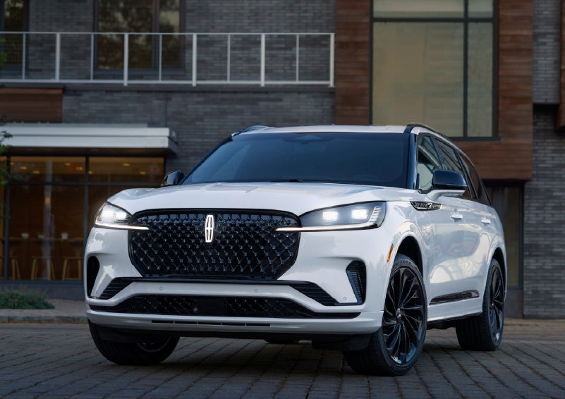 White Lincoln Aviator SUV parked in front of a modern building, shown in a front three-quarter view with LED headlights and black wheels.