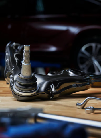 Close-up of a shiny OEM suspension control arm with a ball joint resting on a wooden workbench, with tools and a vehicle in the background.