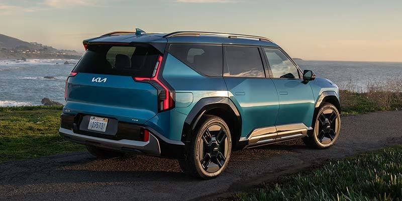 A three-quarter rear view of a metallic blue Kia EV9 electric SUV parked on a coastal path next to the ocean at sunset.