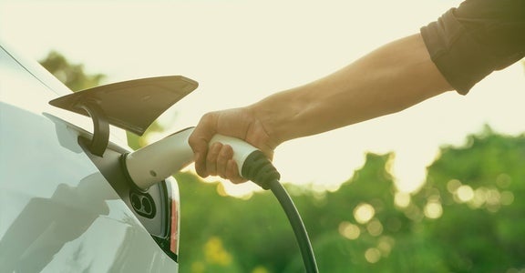 A close-up shot of a person's arm inserting a white charging cable into the charging port of a white electric vehicle, with a blurred green background of trees under sunlight.
