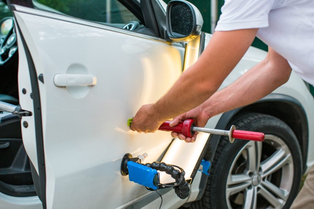 A technician performs Paintless Dent Repair (PDR) on a white car door, using a red slide hammer attached to a glue tab while an LED line board illuminates the panel to highlight the dent.